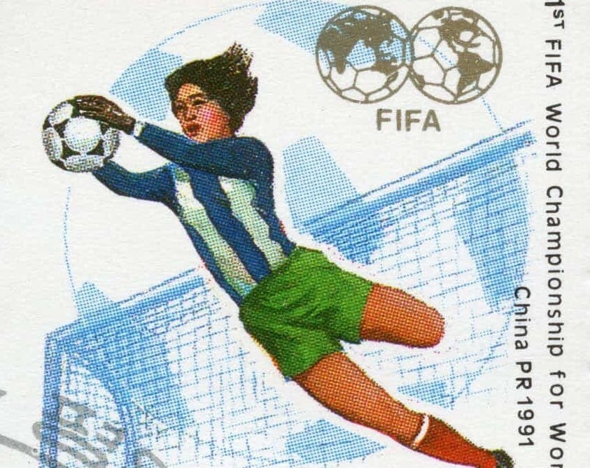 A vintage North Korean stamp depicts a female goalie wearing a blue-striped jersey and green shorts leaping to catch a football A vintage North Korean stamp depicts a female goalie wearing a blue-striped jersey and green shorts leaping to catch a football