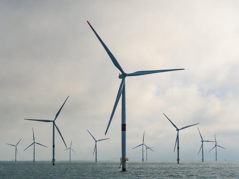 A photograph of an offshore windfarm with turbines in a calm sea under an overcast sky. A photograph of an offshore windfarm with turbines in a calm sea under an overcast sky.