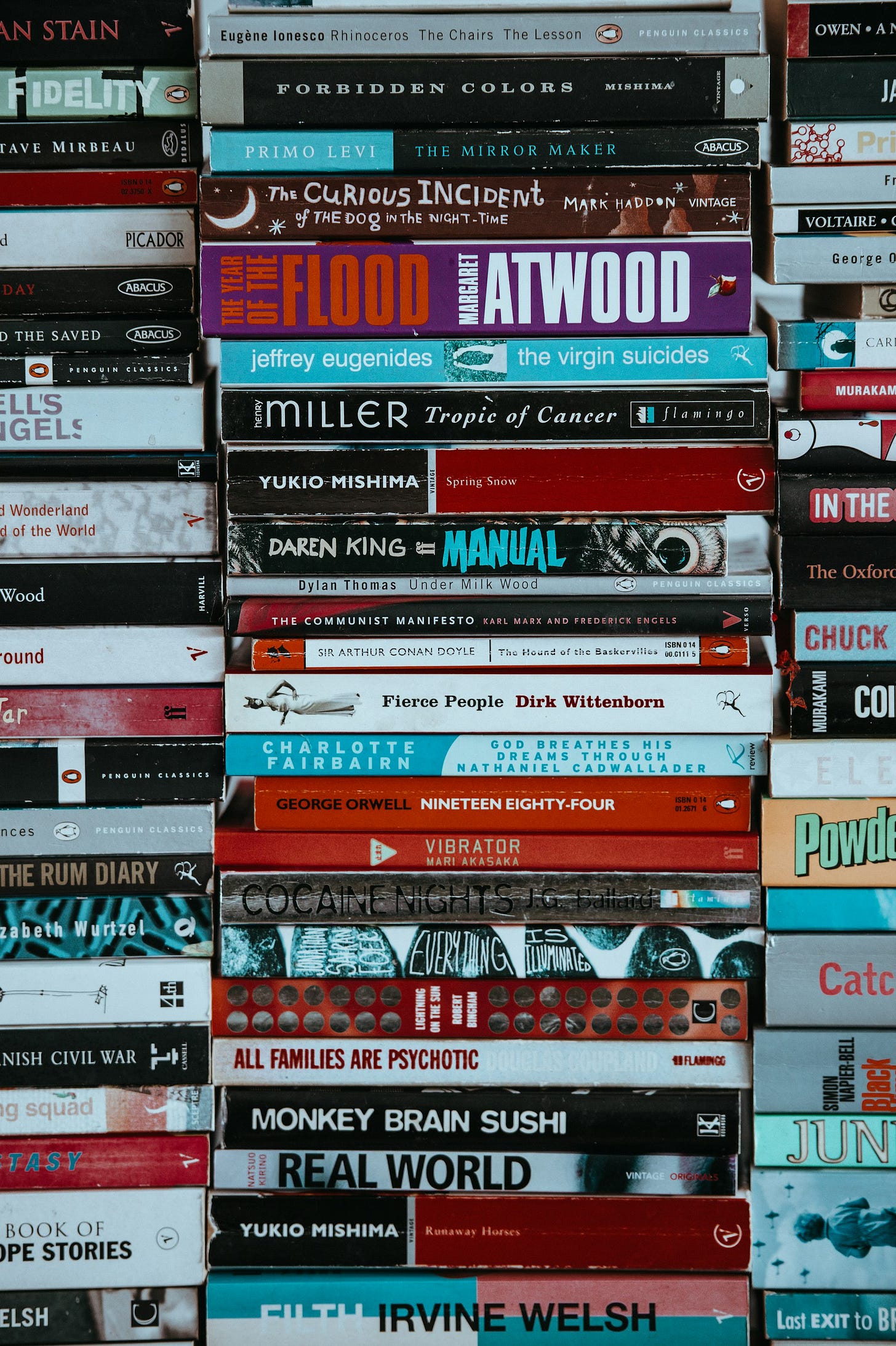 A photo of numerous paperback books stacked in three piles, filling the entire image.