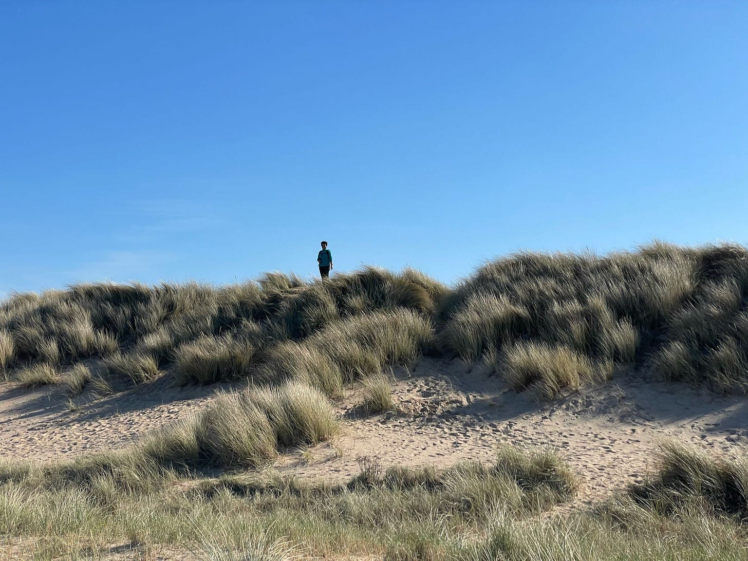 Image shows a woman in the distance standing on the top of sand dunes, wearing a green fleece. There are grasses on the sand dunes and the sky is blue and cloudless behind