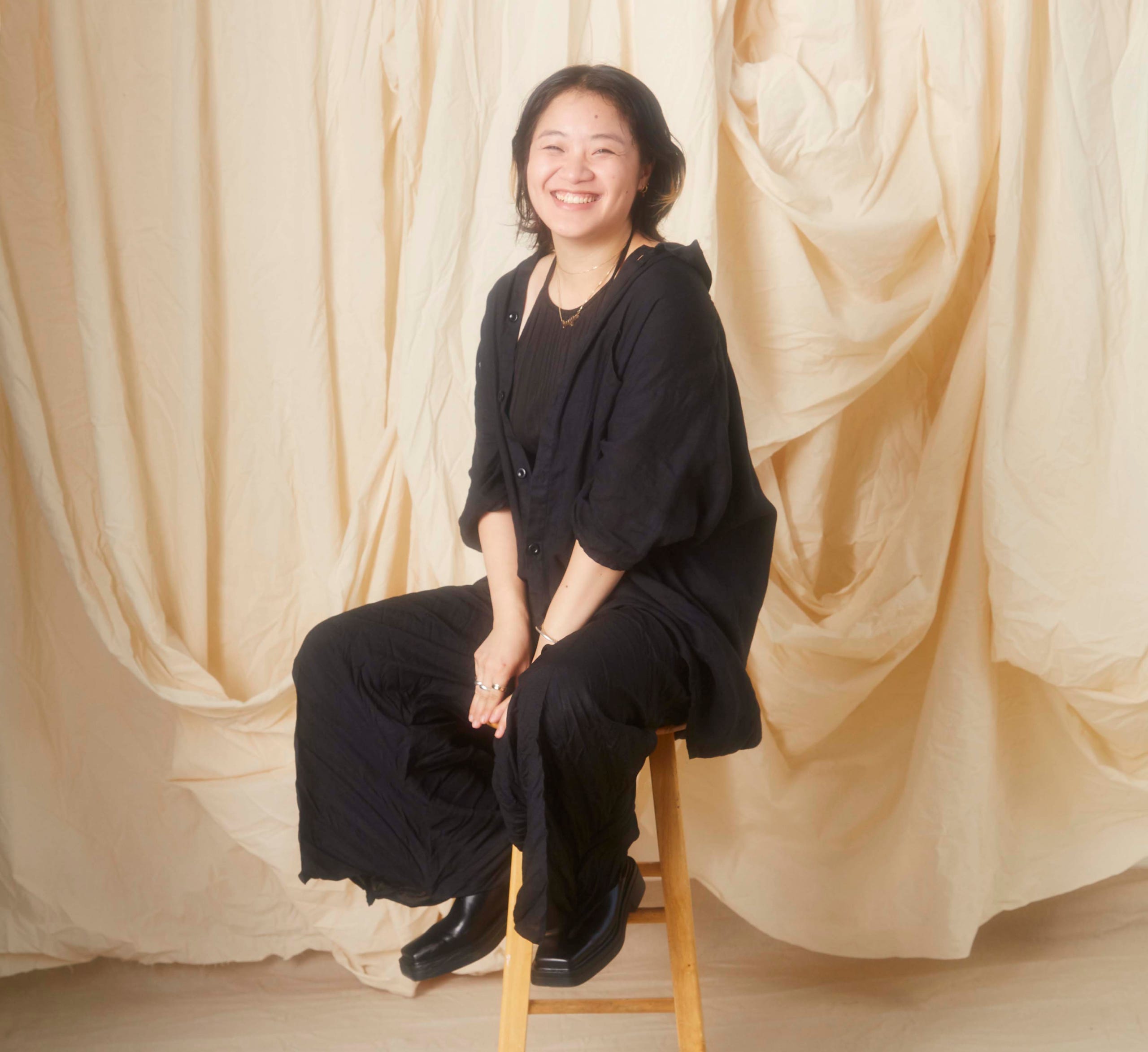 Ka Yee Chans sits on a stool, smiling towards the camera in front of a beige background. Ka Yee Chans sits on a stool, smiling towards the camera in front of a beige background.