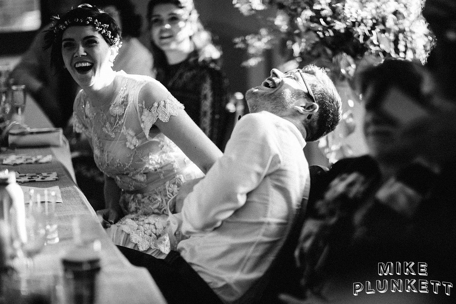 A couple in wedding gear laughing their heads off during the speeches. Janelle has her mouth open in a wide laugh and Tom has his head thrown back