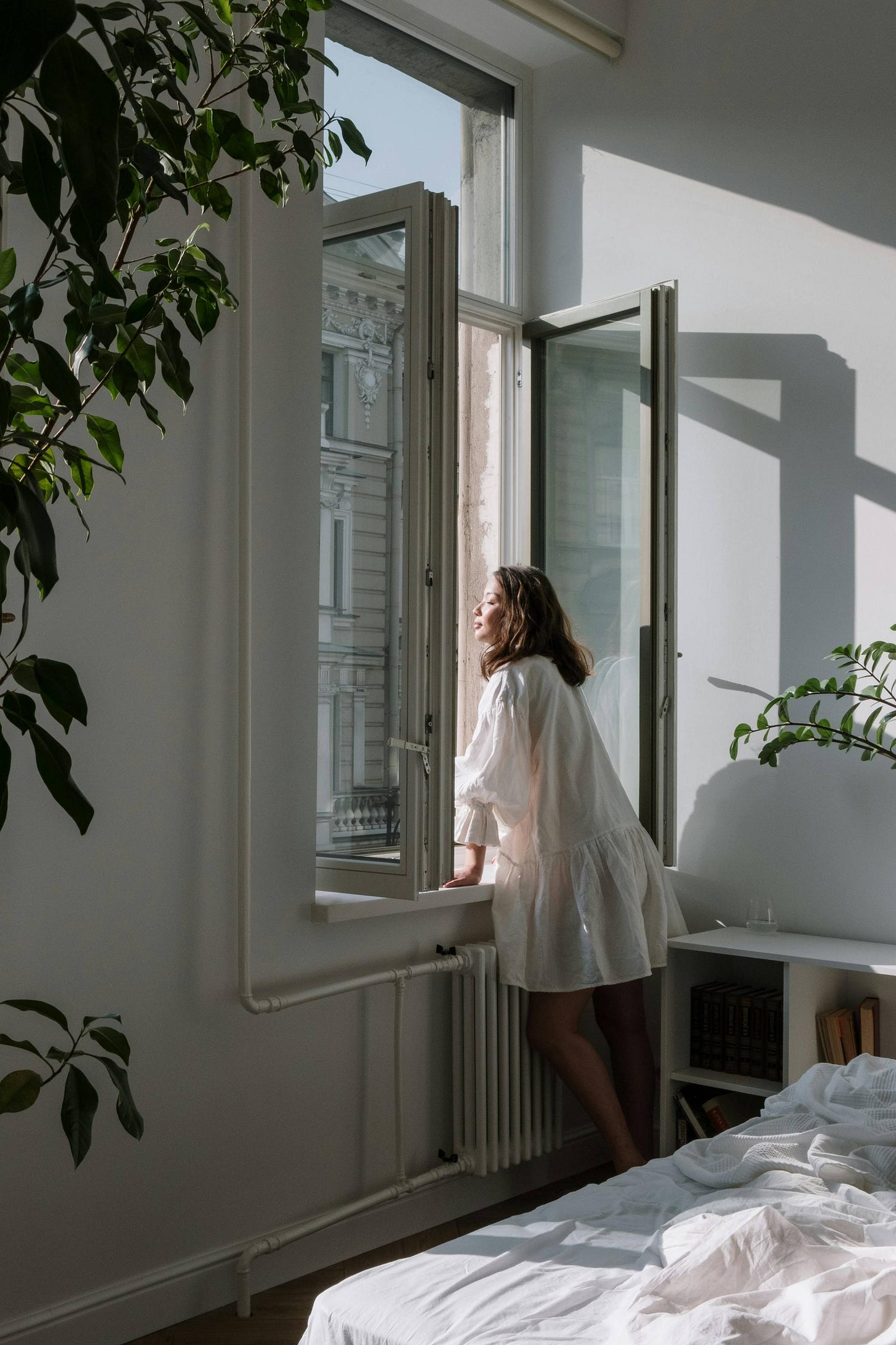  A woman standing by an open window, basking in natural sunlight—representing the importance of morning light exposure in resetting circadian rhythms for better sleep and energy. order of operations for longevity