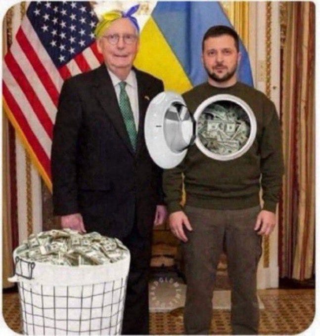 An edited photo depicts Joe Biden dressed in a dark suit with a green tie standing beside Volodymyr Zelenskyy in a green hoodie featuring a circular hole resembling a washing machine drum filled with stacked US dollar bills. They are positioned in front of a blue curtain with American and Ukrainian flags on either side. A white bucket overflowing with loose US dollar bills sits on the floor to the right of Biden. The image has a Giphy watermark in the bottom right corner.