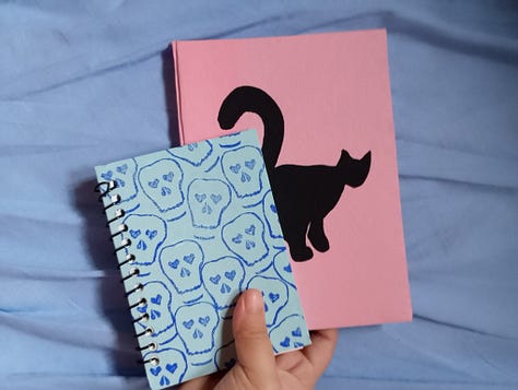Grade de fotos mostra quatro cadernos, um preto, um azul com carimbos azul escuros em formato de caveira, um rosa com um gato preto pintado e um marrom com carimbos azuis de um peixe com pernas
