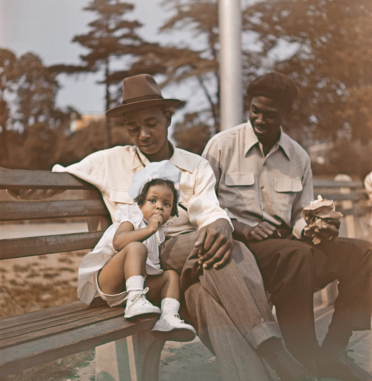 A young girl in a white dress and bonnet sits on a park bench, leaning against a man in a hat and light shirt. Another man beside them smiles while holding food. Trees and benches fill the background. A young girl in a white dress and bonnet sits on a park bench, leaning against a man in a hat and light shirt. Another man beside them smiles while holding food. Trees and benches fill the background.