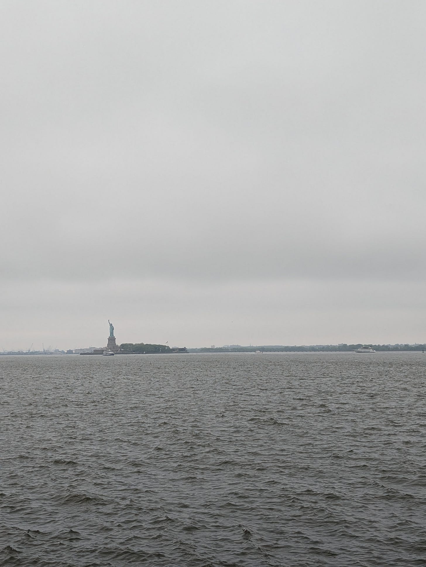 the Statue of Liberty in the distance with gray hazy sky in the background and turbulent river water in the foreground