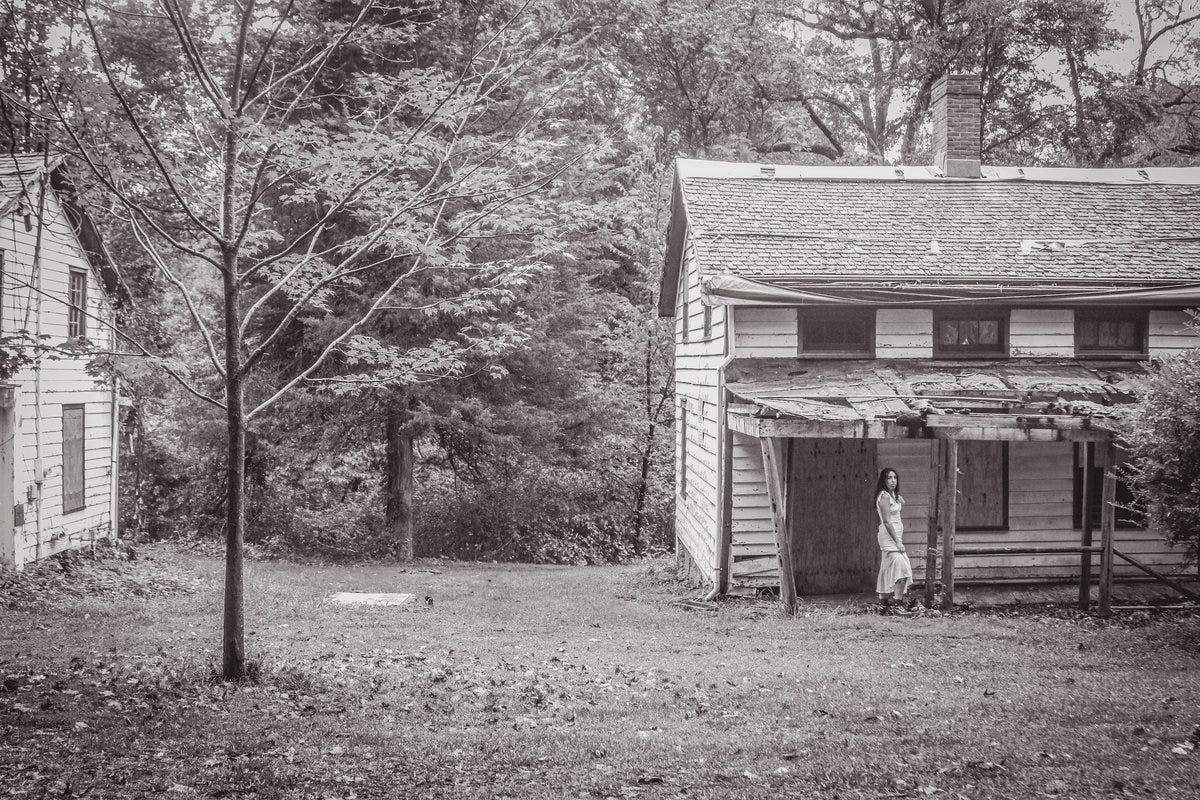 Black and white or grayscale picture of two abandoned homes in a rural setting. There's a larger portion of a home on the right, white and two stories (much like the one slightly visible on the left). This cover photo for Stefanie Carapezza's "Stagefright" by Eli Wagner features Carapezza, in a white dress, standing by the house on the right. A spare tree is in the space between the houses, closer to the one on the left, with woods looming in the background. Black and white or grayscale picture of two abandoned homes in a rural setting. There's a larger portion of a home on the right, white and two stories (much like the one slightly visible on the left). This cover photo for Stefanie Carapezza's "Stagefright" by Eli Wagner features Carapezza, in a white dress, standing by the house on the right. A spare tree is in the space between the houses, closer to the one on the left, with woods looming in the background.