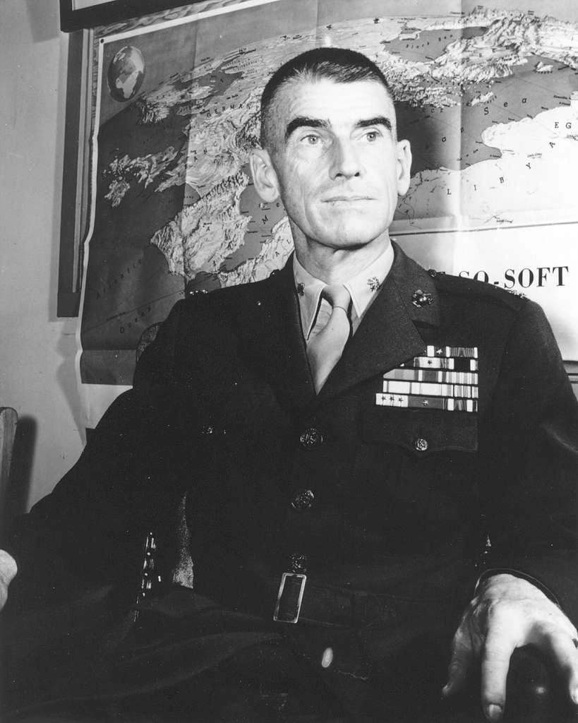 Formal black-and-white portrait of U.S. Marine LtCol Evans F. Carlson in service dress with ribbons and medals, seated in front of a large wall map of the Pacific theater