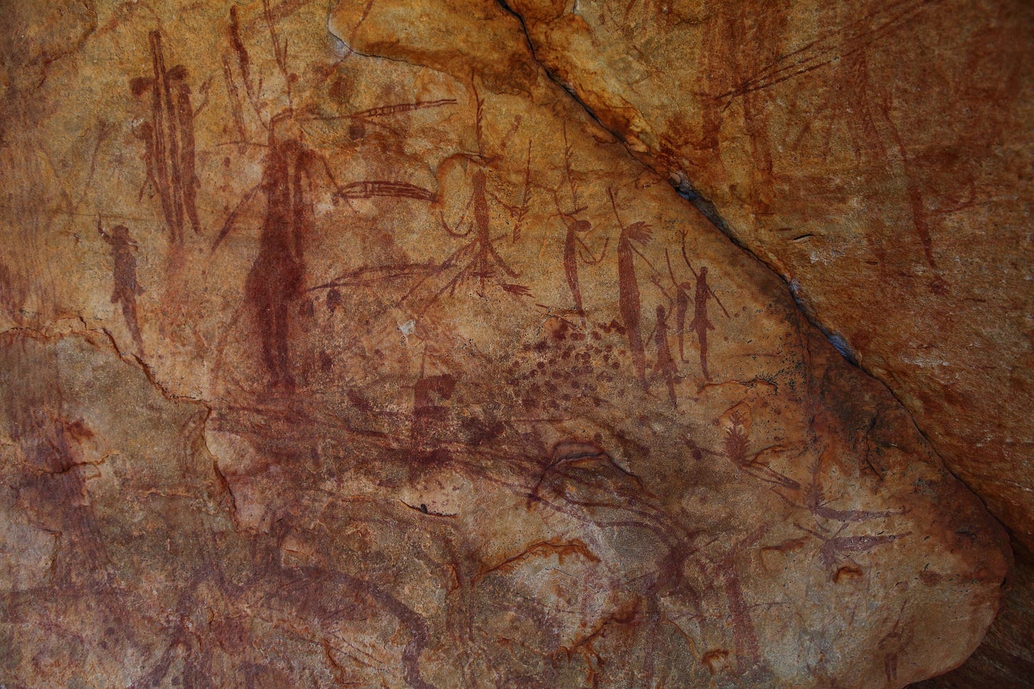 An overhanging sandstone wall in warm ochre tones, covered with slender dark red human figures, some with tassels and headdresses, painted over one another. An overhanging sandstone wall in warm ochre tones, covered with slender dark red human figures, some with tassels and headdresses, painted over one another.