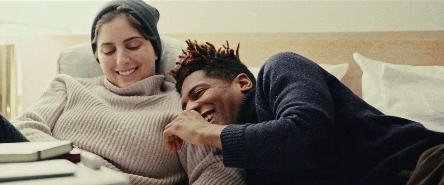 Suleika, a light-skinned woman in a beanie wearing a sweater, lying with Jon, a Black man smiling and wearing a blue sweater. Both are in bed together