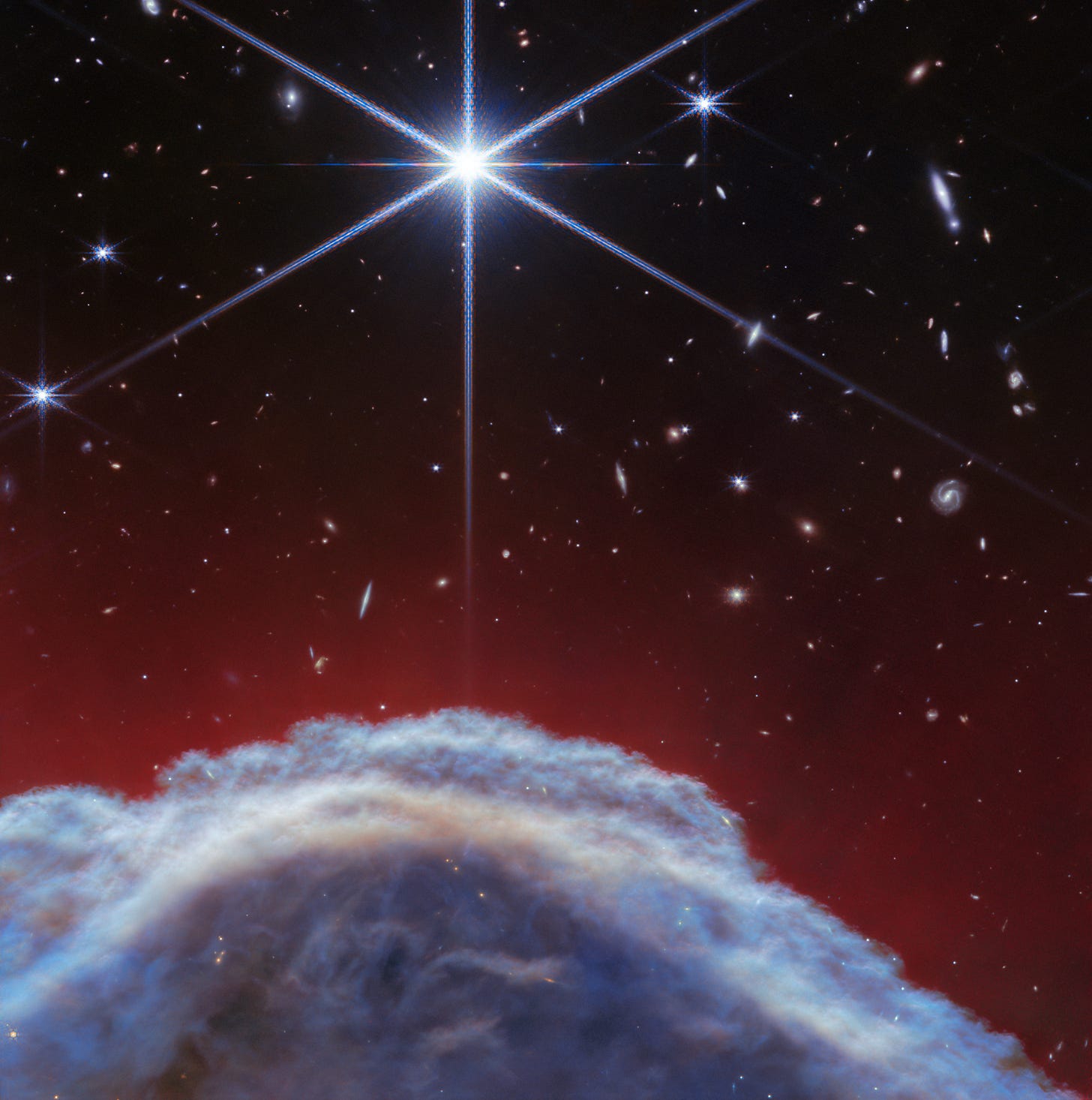A clumpy dome of blueish-gray clouds rises about a third of the way from the bottom. Above it, streaky, translucent red wisps brush upward to about halfway up the image. The top half of the image is the black background of space with one prominent, bright white star with Webb’s 8-point diffraction spikes. Additional stars and galaxies are scattered throughout the image, although very few are seen through the thick clouds at bottom and all are significantly smaller than the largest star. A clumpy dome of blueish-gray clouds rises about a third of the way from the bottom. Above it, streaky, translucent red wisps brush upward to about halfway up the image. The top half of the image is the black background of space with one prominent, bright white star with Webb’s 8-point diffraction spikes. Additional stars and galaxies are scattered throughout the image, although very few are seen through the thick clouds at bottom and all are significantly smaller than the largest star.