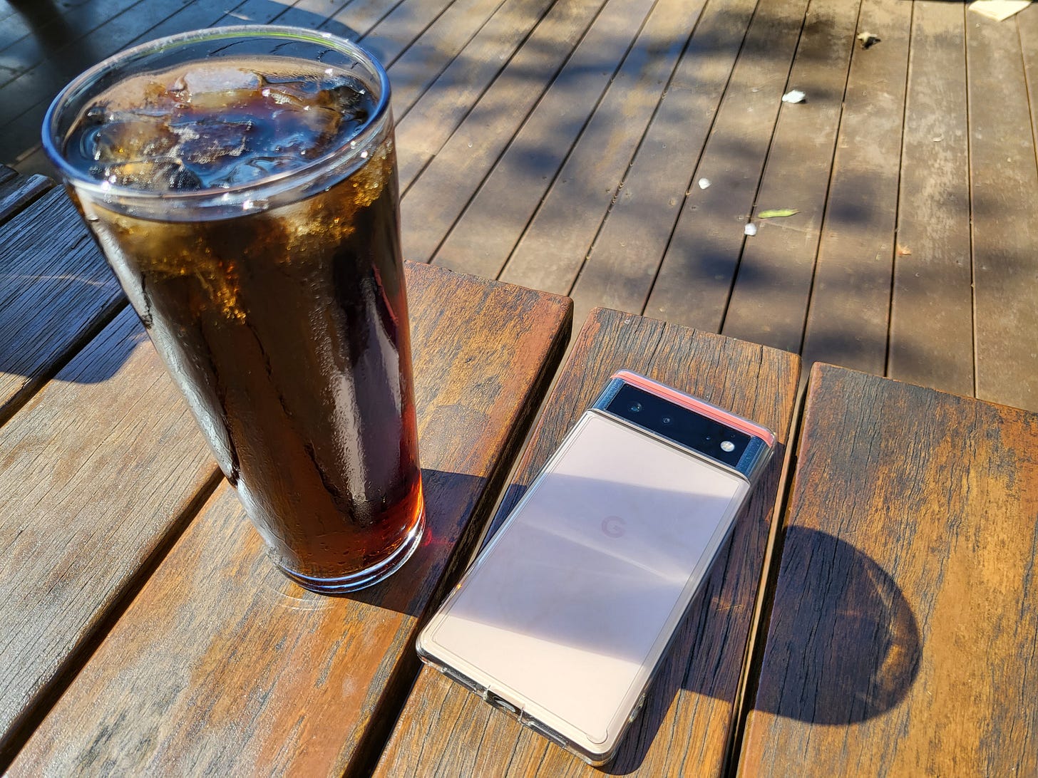 An image of a glass of iced Coke on the left with a phone colored pink-orange and pink-cream in a two-tone manner resting on a wooden table. Also seen are the wooden platform floorboards
