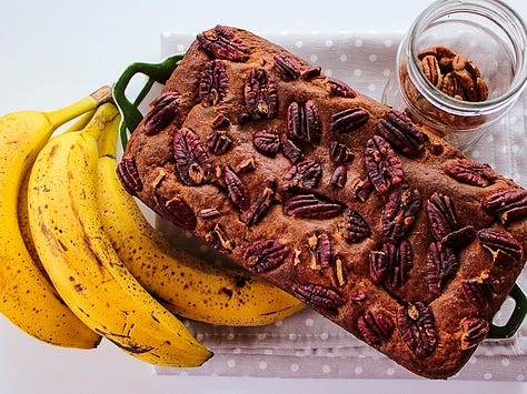 Homemade banana bread topped with pecans, shown whole and sliced to reveal a moist, tender crumb, styled with ripe bananas on a neutral linen background.