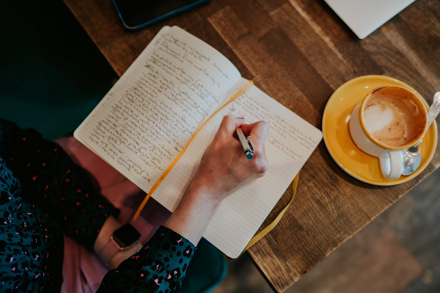 An individual writes in a journal about wellbeing sat at a table next to a cappuccino in a yellow cup. 