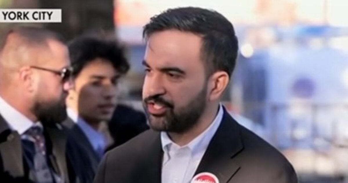 Man speaking at a public event in New York City, wearing a campaign button, surrounded by a group of people in the background.