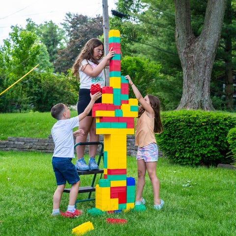 Children building a colorful, tall tower with Biggo Blocks in a backyard, using teamwork and creativity to stack the large building blocks as high as possible.
