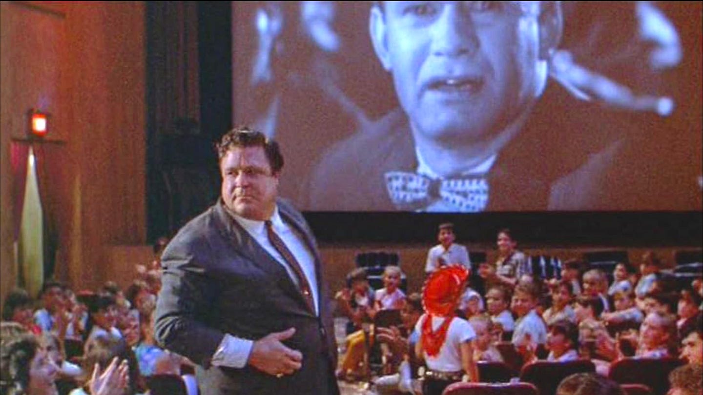 Lawrence Woolsey (John Goodman) stands in front of a packed audience during a screening of Mant!, a giant face projected behind him on the theater screen. Lawrence Woolsey (John Goodman) stands in front of a packed audience during a screening of Mant!, a giant face projected behind him on the theater screen.
