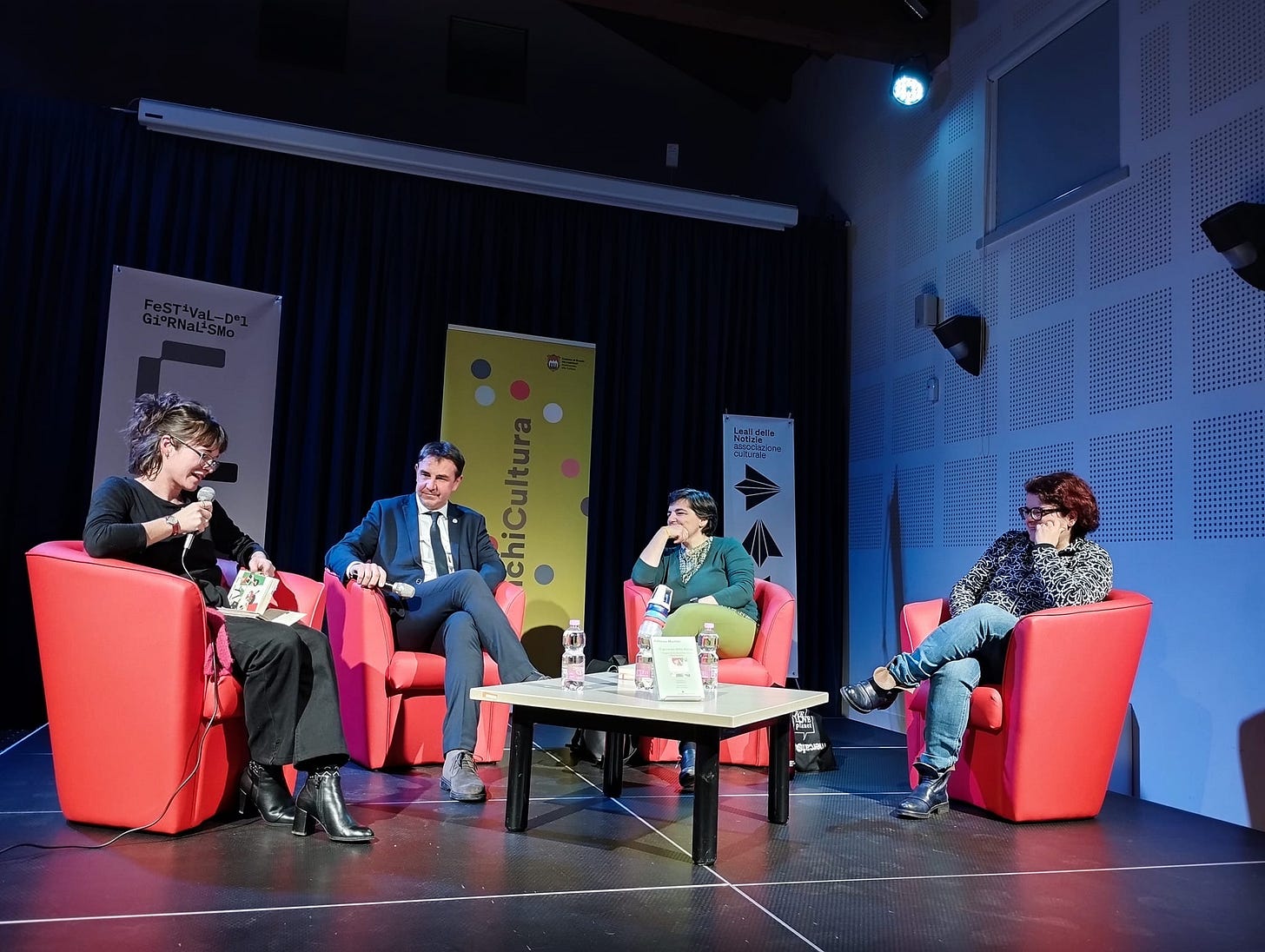 Quattro persone sedute su un palco, tutte su una poltroncina rossa. Una delle persone, quella più a sinistra, ha il microfono in mano e sta parlando.