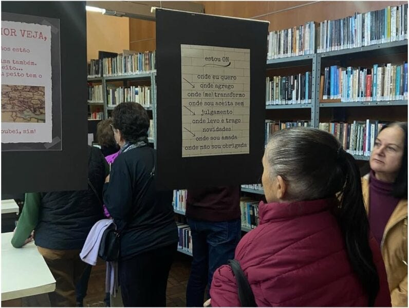 #descrevemospravocê  A imagem mostra o interior de uma biblioteca durante uma exposição textual. Várias pessoas, em sua maioria mulheres adultas e idosas, observam painéis pendurados no centro da sala, que trazem textos impressos em folhas fixadas sobre placas pretas. Ao fundo, estantes metálicas estão repletas de livros organizados. A iluminação do espaço é suave e o ambiente é acolhedor.  No centro da imagem, um dos painéis em destaque exibe um texto em letras digitadas sobre uma folha pautada, com os seguintes dizeres: “estou ON onde eu quero onde agrego onde (me) transformo onde sou aceita sem julgamento onde levo e trago novidades onde sou amada onde não sou obrigada”.  À frente deste painel, uma mulher de cabelos grisalhos presos em rabo de cavalo, vestindo um casaco acolchoado vinho, observa atenta. Outras pessoas, também de costas ou perfil, circulam e leem os materiais expostos. A imagem transmite uma atmosfera de escuta, reflexão e valorização da palavra em um espaço de leitura e convivência.