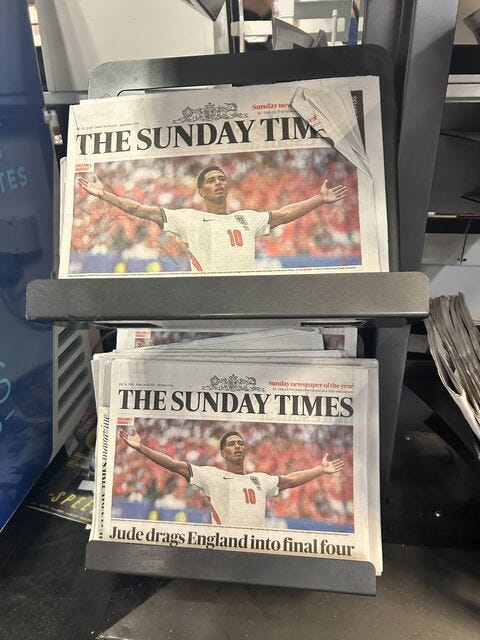 A photograph of copies of The Sunday Times newspaper stacked on a retail display stand, showing the front-page headline 'Jude drags England into final four' with a large picture of football player Jude Bellingham celebrating with open arms. A photograph of copies of The Sunday Times newspaper stacked on a retail display stand, showing the front-page headline 'Jude drags England into final four' with a large picture of football player Jude Bellingham celebrating with open arms.