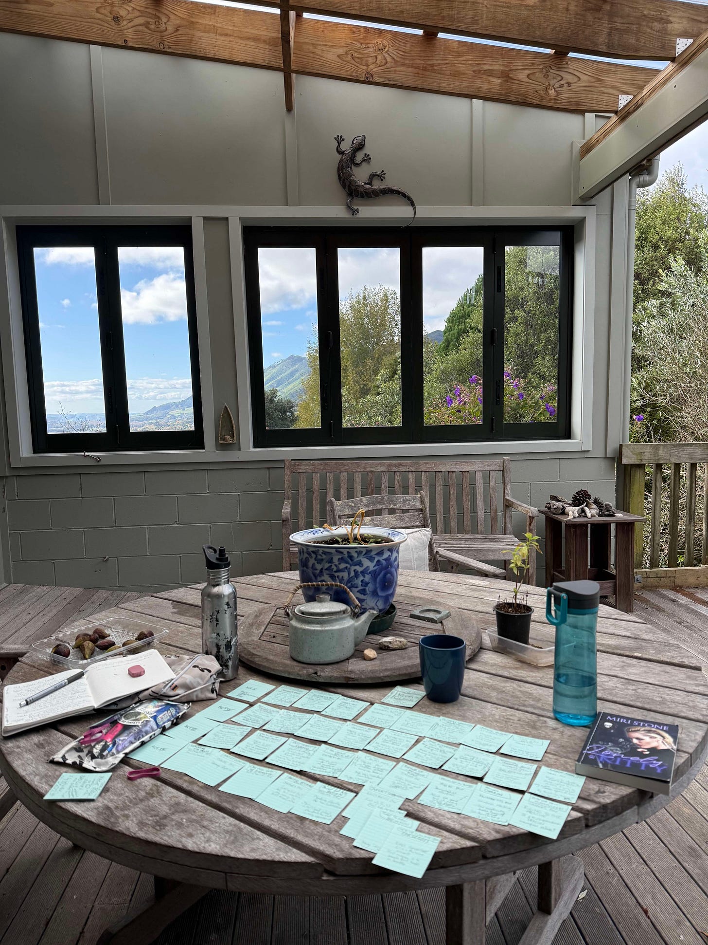 Picture of Miri's writing set up on back deck of her family home in New Zealand with her beat cards spread out on the table and a gorgeous rural view in the background