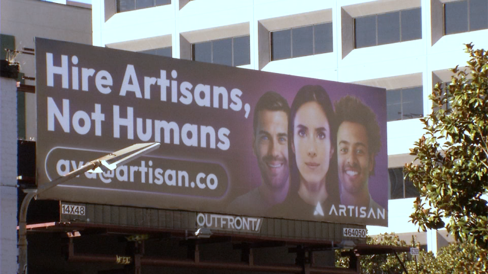 Billboard on a city street reading “Hire Artisans, Not Humans” above the website “artisan.co,” featuring three portrait photos of three adults on a dark gradient background, with an office building and trees behind the sign.