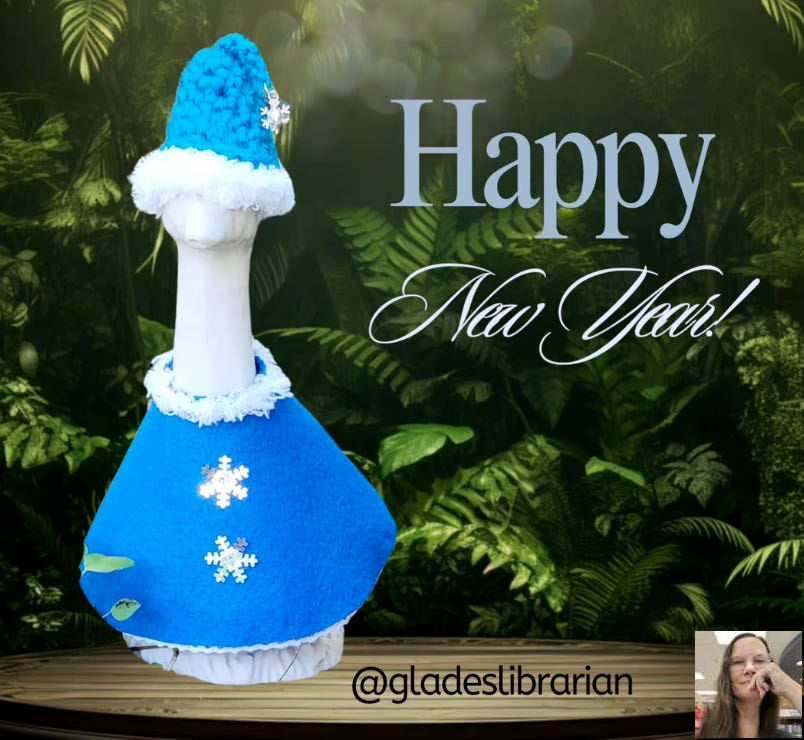 Image of all white 3D-printed 7" desk goose dressed in a sky blue felt sweater with two snowflake-shaped silver buttons and a white furry round neckline. Also wearing a sky blue pointed hat with white fur edging along the forehead. A small silver snowflake jewelry finding acts as a tassel. Text reads "Happy New Year!" and "@gladeslibrarian".