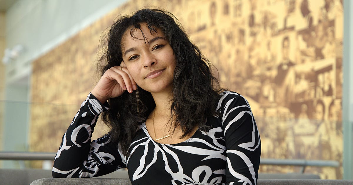 Nia Pearl wearing a black and white top, head leaning slightly on her hand, in front of a sepia-toned mural wall