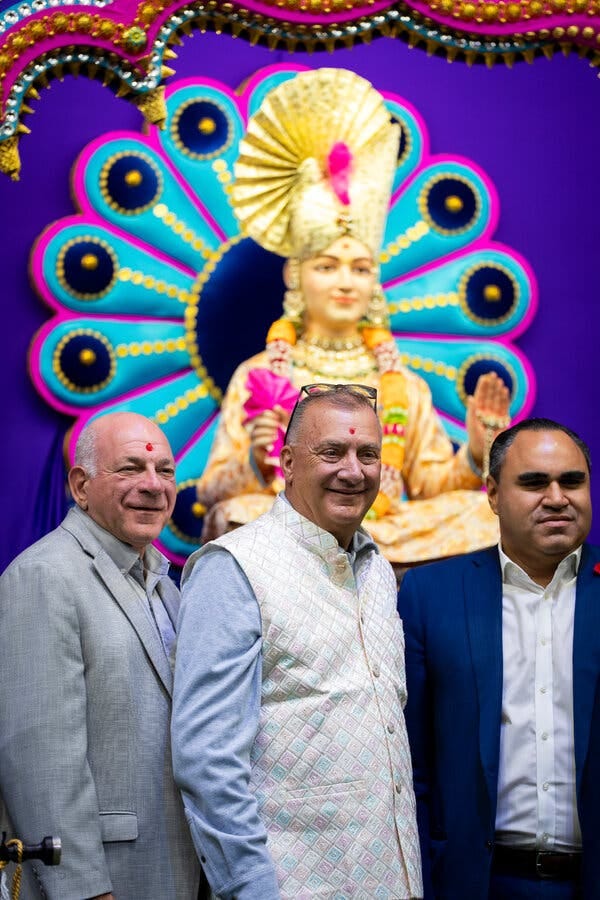 In front of a golden Hindu statue in front of a brightly painted background of blues and purples stand three men, including Mayor Gonnelli; two of them, including the mayor, have red bindi on their foreheads.