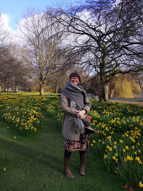 Three photos of me in St. James' Park in February 2020. I am standing surrounded by daffodils, and whilst I am rugged up in my grey coat and blanket scarf, a floral skirt peeks out from underneath and I am clearly embracing the new season. The background includes three big tree still in Winter mode but a small patch of blue sky is peeking through the clouds. Photos taken by S-