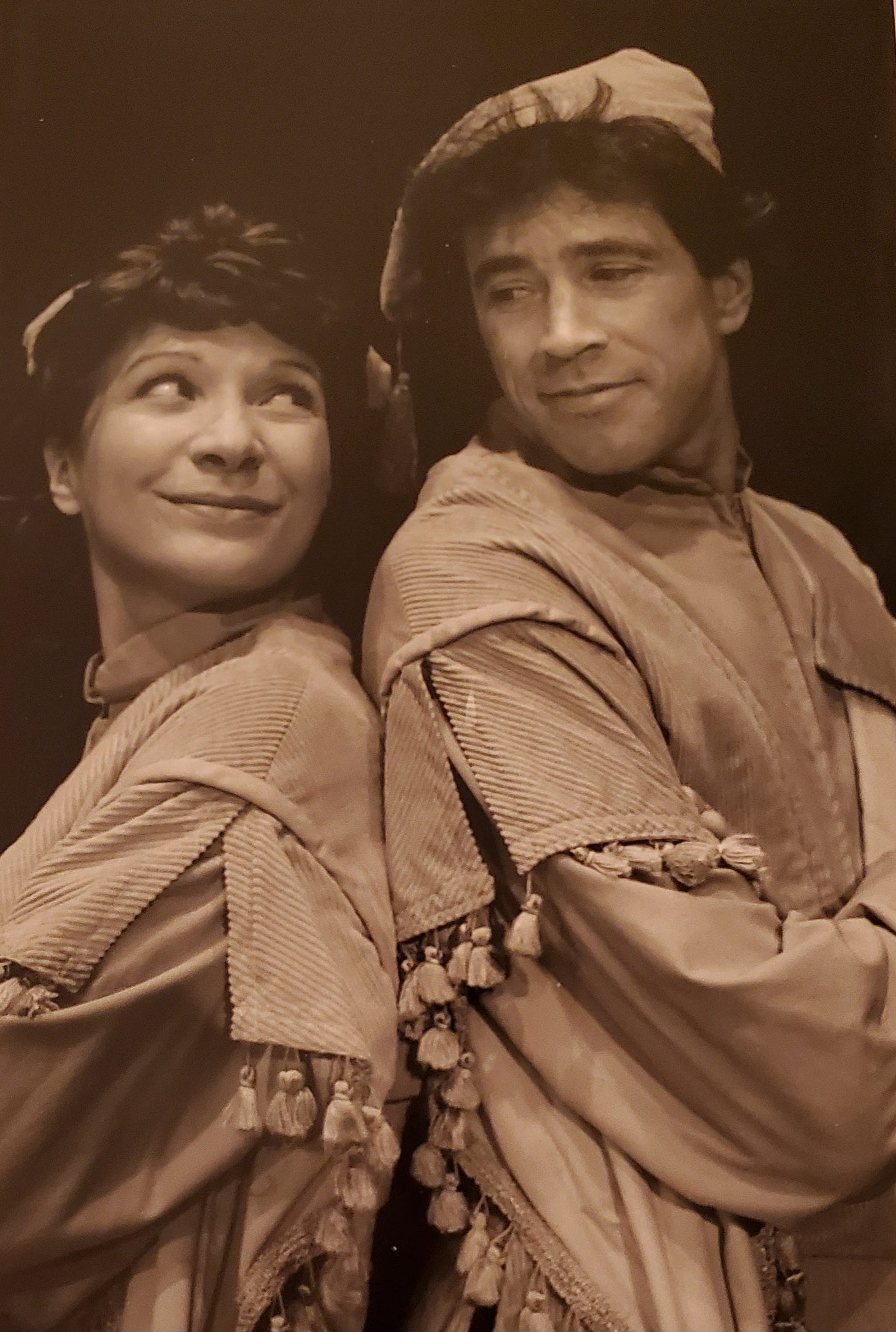 A white woman & man in Elizabethan costume stand back to back, smiling at each other over their shoulders