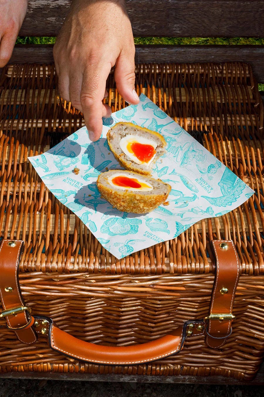A hand reaching for a cut open scotch egg that is on top of a Fortnum's hamper.