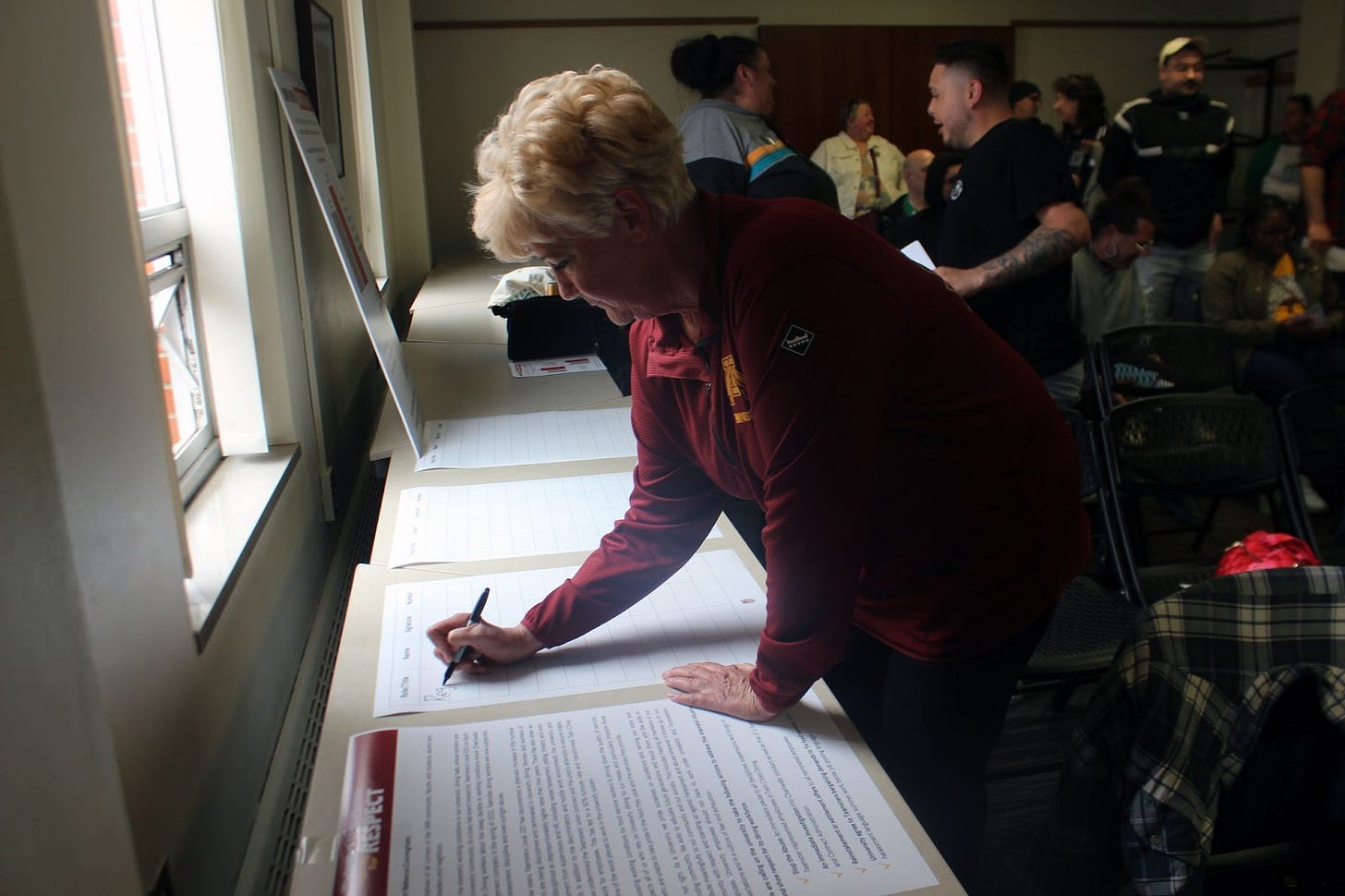 a blonde haired woman wearing a maroon quarter zip sweater leans down a table to sign a petition a blonde haired woman wearing a maroon quarter zip sweater leans down a table to sign a petition