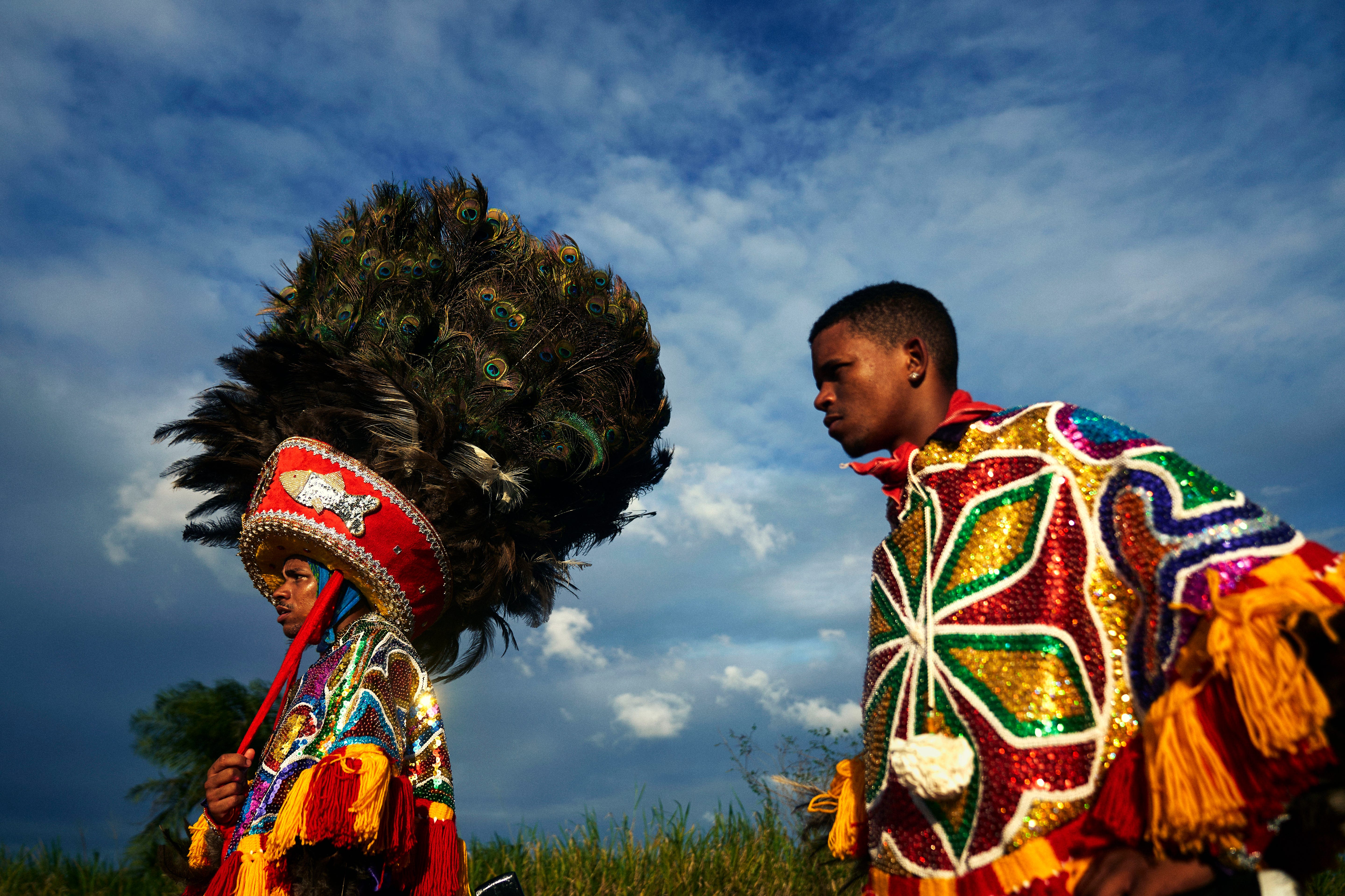 Two people wear vibrant, sequined costumes; one has a large feathered headdress. They stand outdoors under a dramatic, cloudy sky with green grass in the background. Two people wear vibrant, sequined costumes; one has a large feathered headdress. They stand outdoors under a dramatic, cloudy sky with green grass in the background.