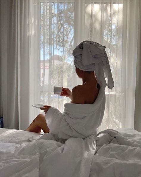 This may contain: a woman sitting on a bed with a towel wrapped around her head and holding a cup