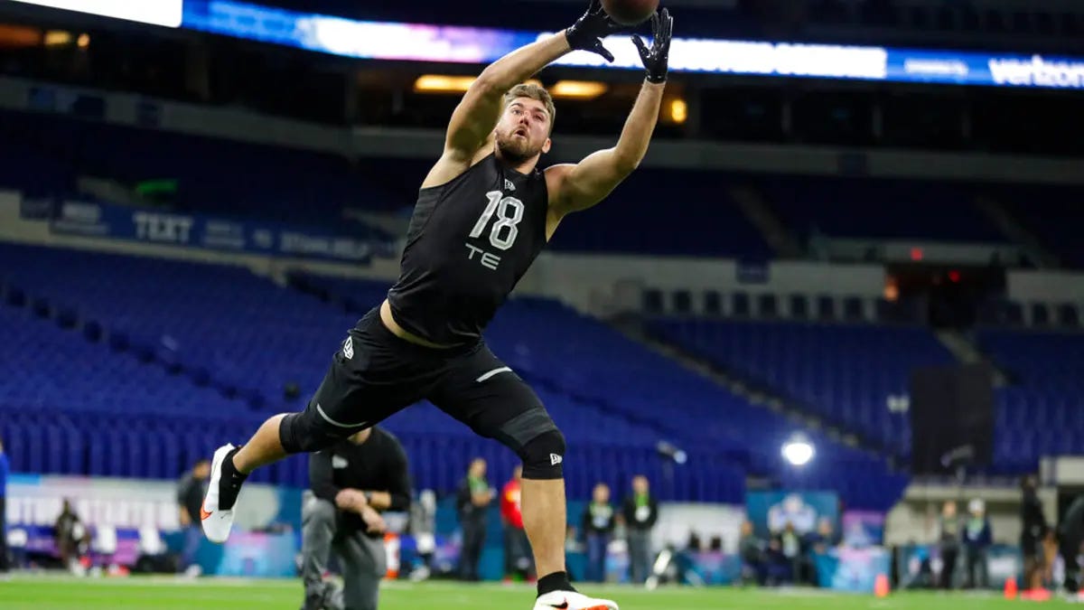 a tight end running on an empty stadium in an extended position about to catch a football at the top of their reach