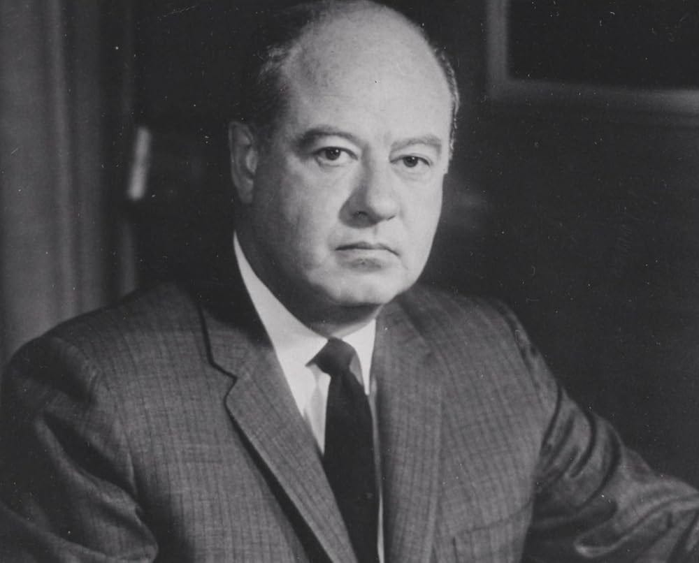 Robert Maheu appears to be sitting in an office, but he's too close to say for sure. He's wearing a dark gray suit and a black tie. His body is turned to three-quarters profile, but his face is turned toward the camera. He doesn't look happy.