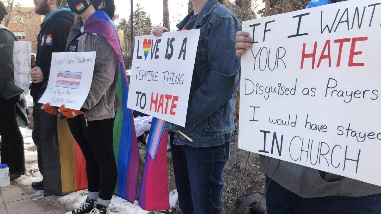 Protesters, many clad in rainbow garb or capes, hold up signs. One visible sign reads: "Love is a terrible thing to hate." Another reads, "If I wanted your hate disguised as prayers, I would have stayed in church." Protesters, many clad in rainbow garb or capes, hold up signs. One visible sign reads: "Love is a terrible thing to hate." Another reads, "If I wanted your hate disguised as prayers, I would have stayed in church."