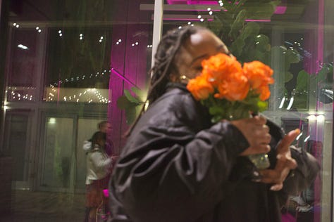 A cinematic rooftop night sequence captured in low light: orange roses glowing under warm interior lamps, reflections across glass obscuring a woman’s face as she lifts the bouquet, illuminated concrete stairs rising into shadow, a white poodle leaning from a car window through city light flares, a motion-blurred skyline beneath a crescent moon, and a circular fire pit burning at the center of a table while hands gather around it. The composition prioritizes atmosphere, reflection, and negative space over direct portraiture.