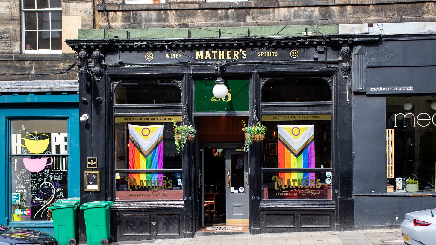 The black frontage of a pub on a busy street. The sign is in gold writing. Two rainbow pride flags hang in the windows.