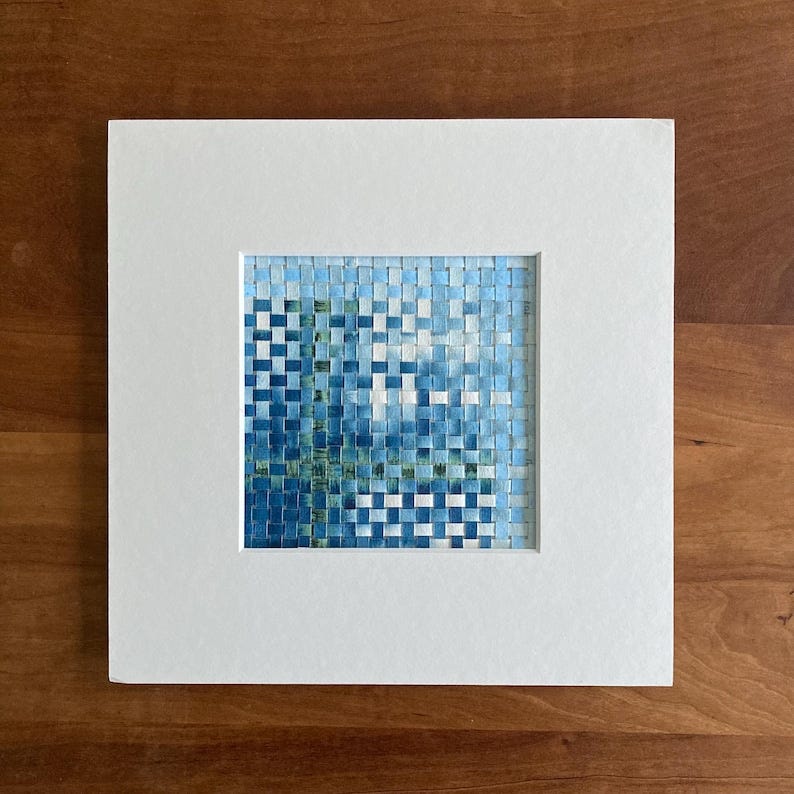 May include: A framed artwork with a blue and green woven pattern. The artwork is framed with a white mat and is displayed on a wooden surface.