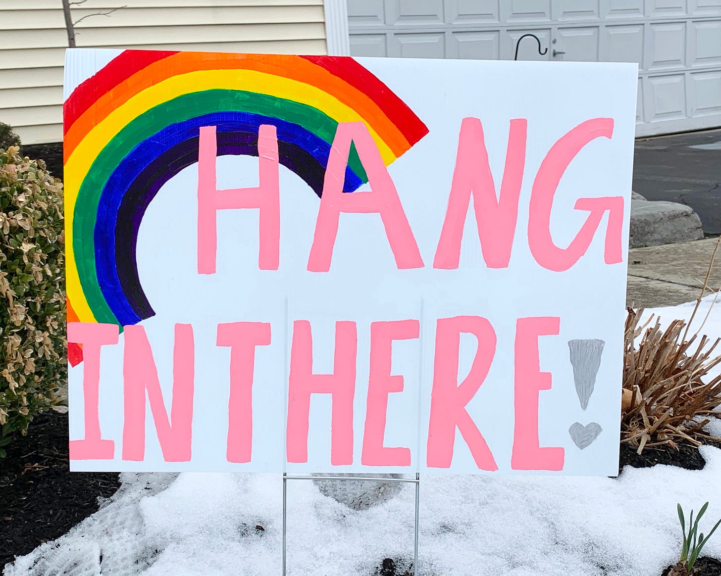 a yard sign with a rainbow painted in the left corner and the words "hang in there" painted in pink letters. 