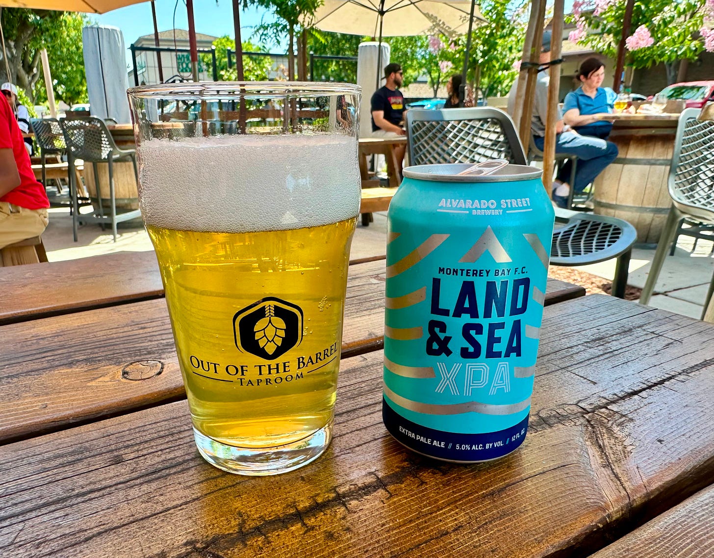A glass of beer next to the can it came from. A beer garden patio in the background A glass of beer next to the can it came from. A beer garden patio in the background