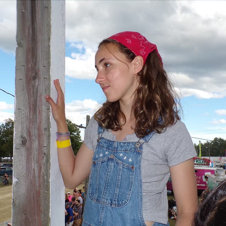 A young woman with curly hair wearing a pink bandana and denim overalls, standing near a wooden post, with a thoughtful expression against a backdrop of a cloudy sky and a crowd in the distance.