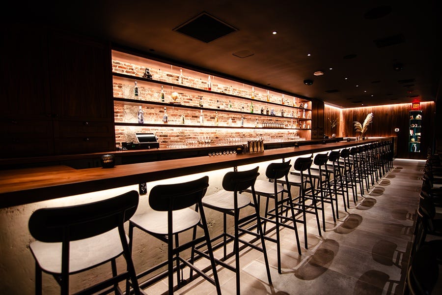 A long, narrow walnut bar is dimly lit. There's exposed brick behind the back bar.