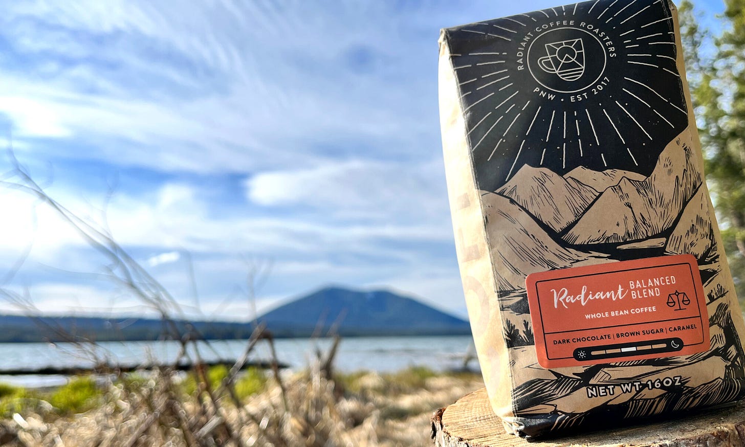 A blurred mountain against a blue sky brushed with white clouds in the background. In the front, right, of the frame is a brown coffee bag with a mountainscape drawn across the front over the orange name label. A blurred mountain against a blue sky brushed with white clouds in the background. In the front, right, of the frame is a brown coffee bag with a mountainscape drawn across the front over the orange name label.