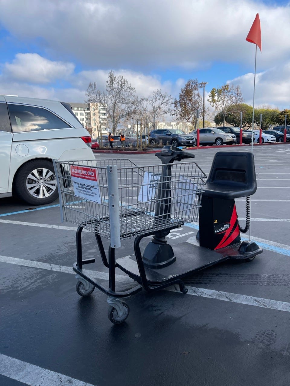 A motorized mobility shopping cart with a wire basket, black seat, and tall orange safety flag parked in a wet grocery-store parking lot under a partly cloudy sky, with a sign on the basket reading "IN-STORE USE ONLY."