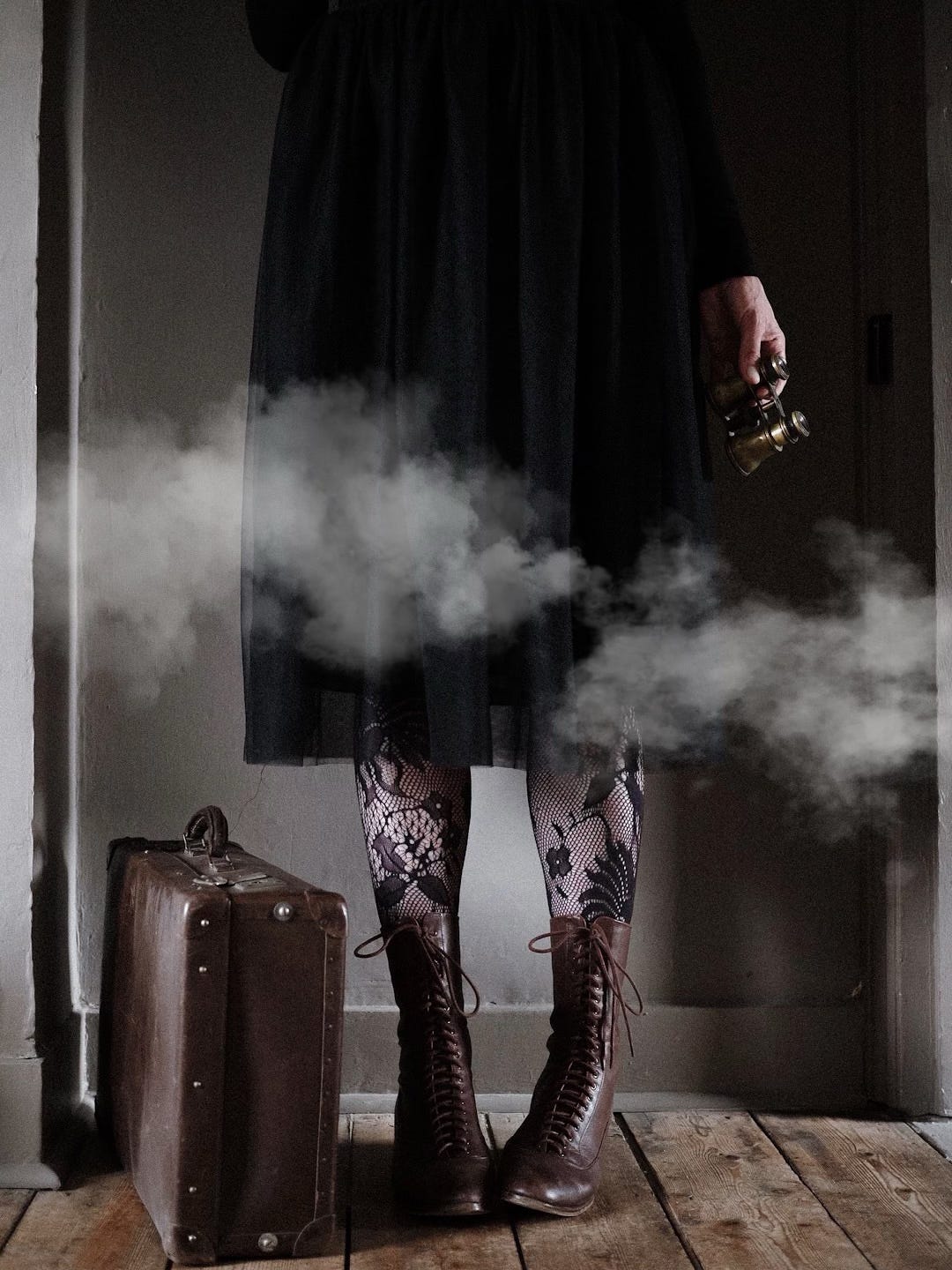 A woman seen from the waist down is wearing a tulle skirt, lace tights and lace-up leather boots, and holding gold binoculars. There are clouds drifting by and a suitcase by her feet.