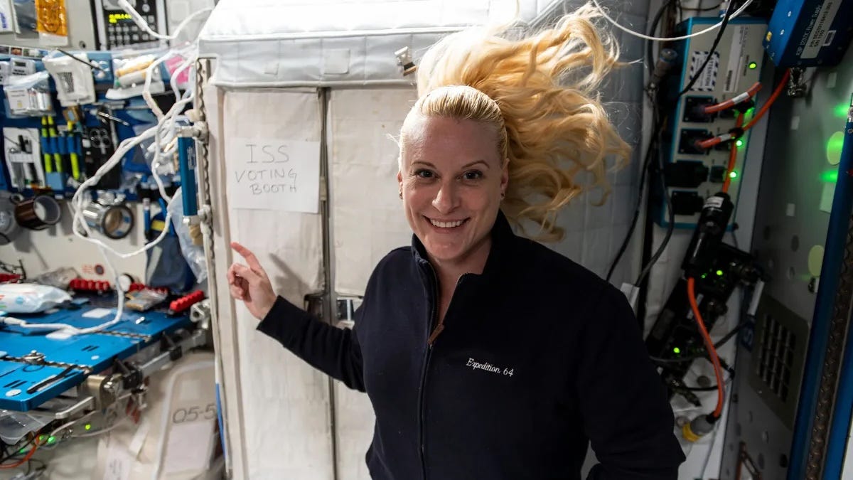 A photo of a woman inside the International Space Station, her ponytailed hair floating in zero-g. She is smiling at the camera and pointing to a handwritten sign taped to a large cabinet behind her, which reads "ISS Voting Booth" A photo of a woman inside the International Space Station, her ponytailed hair floating in zero-g. She is smiling at the camera and pointing to a handwritten sign taped to a large cabinet behind her, which reads "ISS Voting Booth"